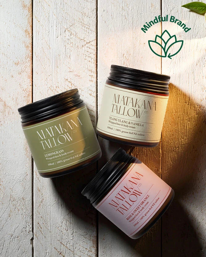 Three jars of Matakana Tallow body butters arranged diagonally on a rustic white-washed wooden background. The products include Lemon Grass, Blanc Blanc & Vanilla, and Peppermint Spice, each in a dark glass jar with a black lid and a cream-colored label.