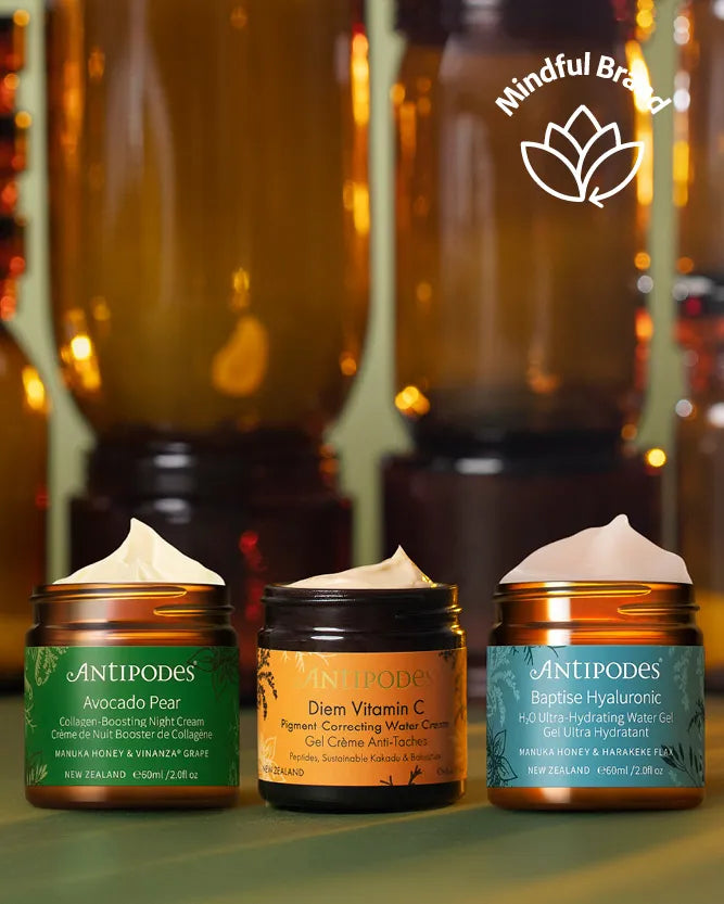 Three Antipodes skincare jars arranged in a row on a green surface with a blurred amber background. The left jar is green and labeled Avocado Pear, the center jar is orange and labeled Diam Vitamin C, and the right jar is amber-brown and labeled Baptise Hyaluronic.