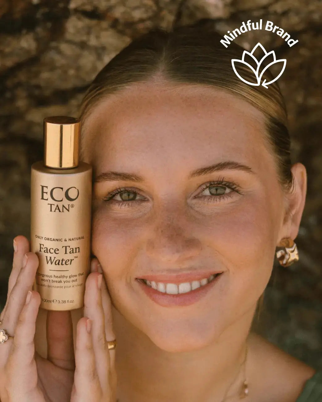 A woman holds a bottle of ECO TAN Face Tan Water next to her face. The bottle is a warm beige color with a metallic gold cap.