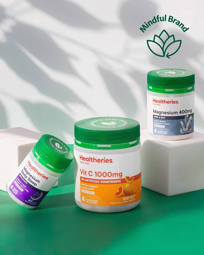Three Healtheries supplement bottles arranged on white blocks against a white background with leaf shadows. The center bottle is a white jar with a green lid and orange label for Vit C 1000mg; the left bottle is a smaller white bottle with a green lid and purple label for Magnesium; the right bottle is a white jar with a green lid and blue label for Magnesium 400mg.