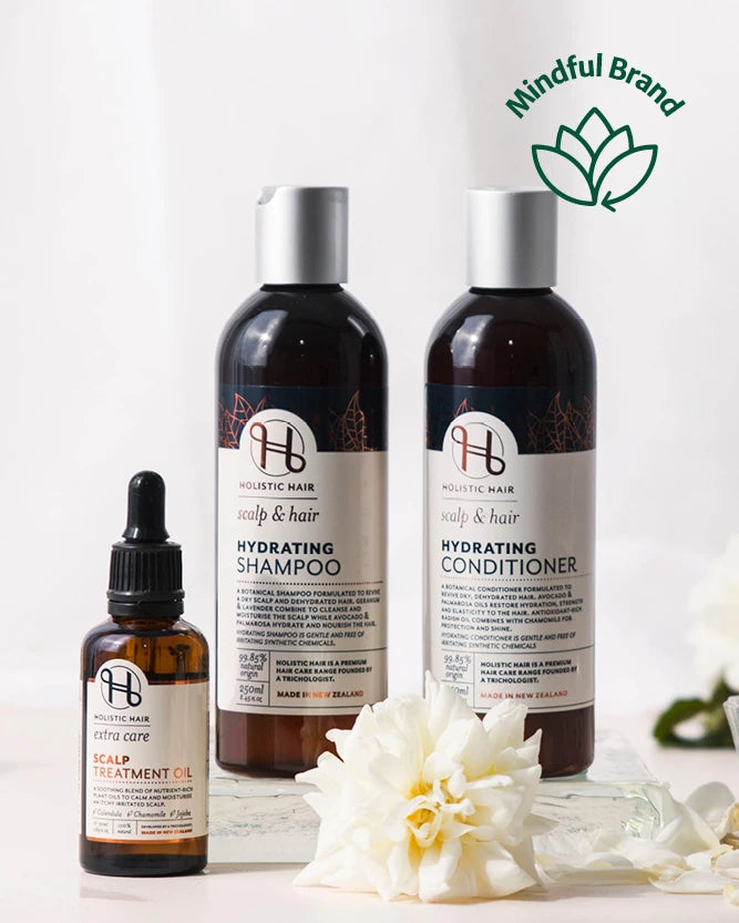 Three hair care products from Holistic Hair are arranged on a white surface: two large dark bottles labeled Hydrating Shampoo and Hydrating Conditioner with silver caps, and one smaller amber dropper bottle labeled Healing Treatment Oil. White flowers are placed in front, and a green 'Mindful Brand' logo appears in the top right corner.
