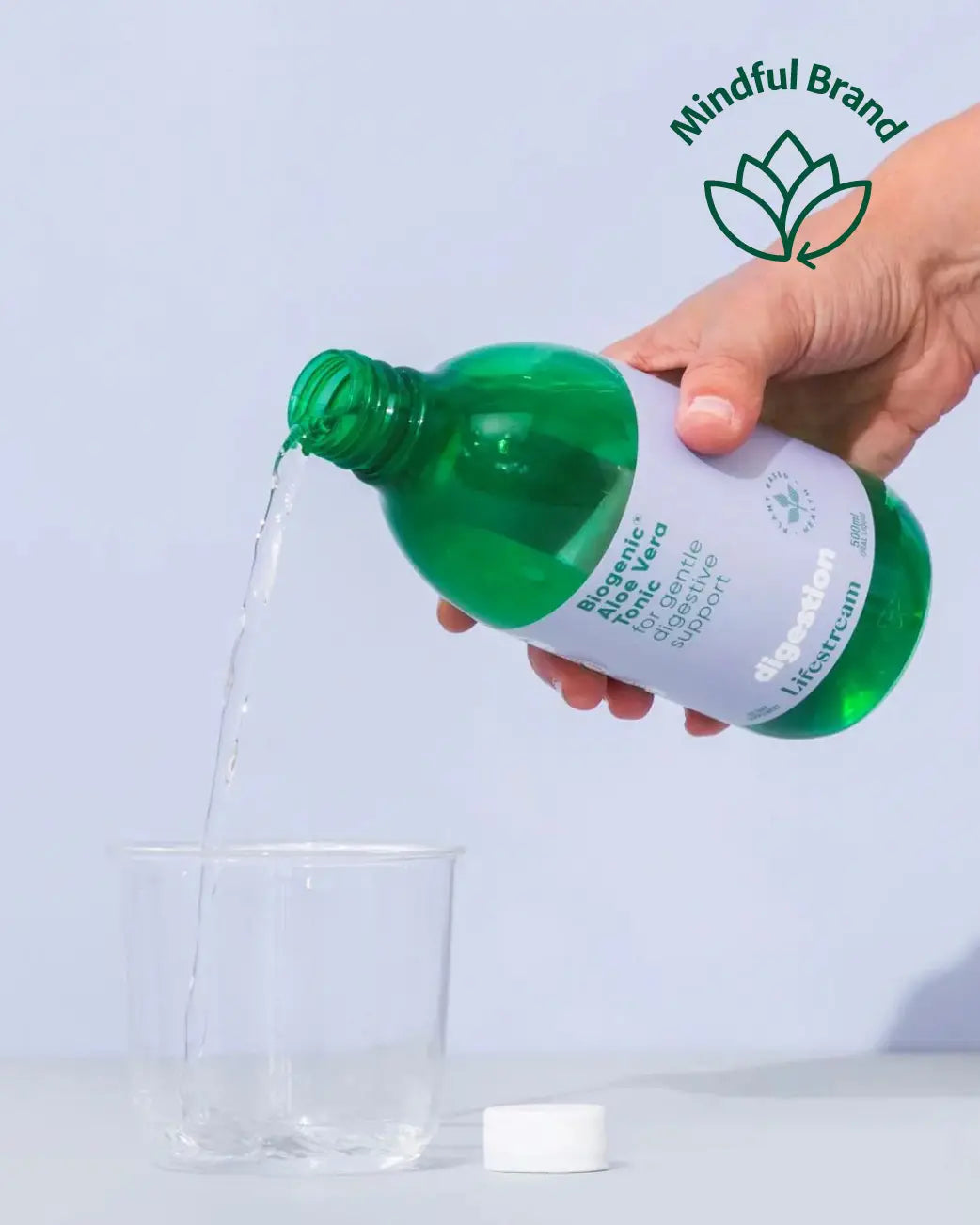 A hand holds a green bottle of Mindful Brand Bioorganic Micelle Cleanser, pouring clear liquid into a clear plastic cup against a light blue background.
