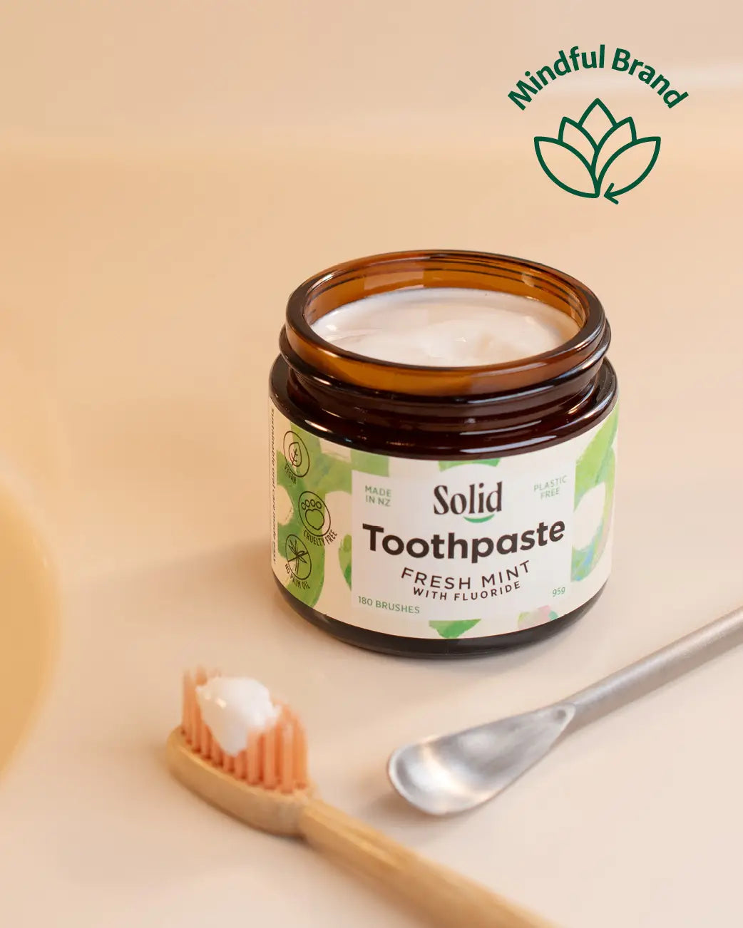 Solid Toothpaste in a clear amber jar with a white and green label, shown on a warm beige background with a wooden toothbrush and metal spoon holding toothpaste. A 'Mindful Brand' logo is visible in the top right.