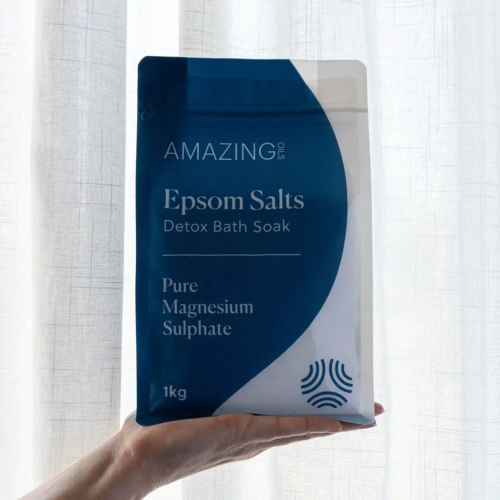 Hand holding a package of Amazing You Epsom Salts Detox Bath Soak against a light curtain background