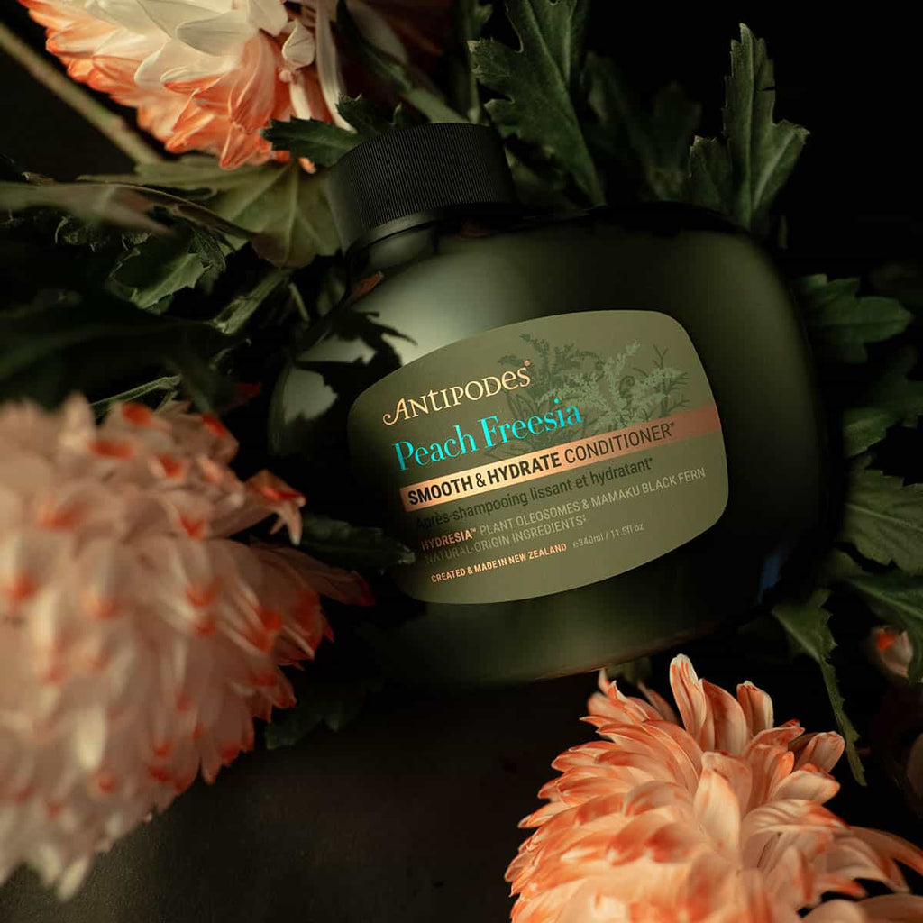 Bottle of Anipodes Peach Freesia conditioner surrounded by flowers on a dark background.