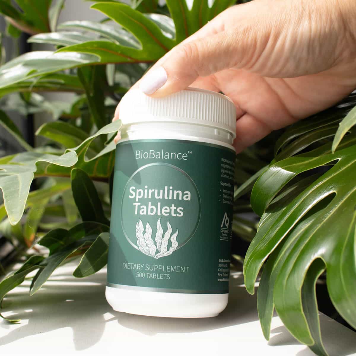 BioBalance Spirulina 500 Tablets bottle held by a hand with green leaves in the background.