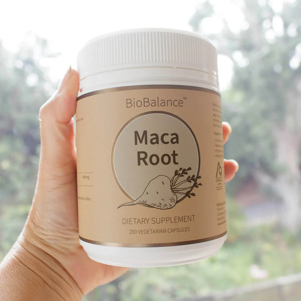 Hand holding a BioBalance Maca Root dietary supplement container with a blurred outdoor background.