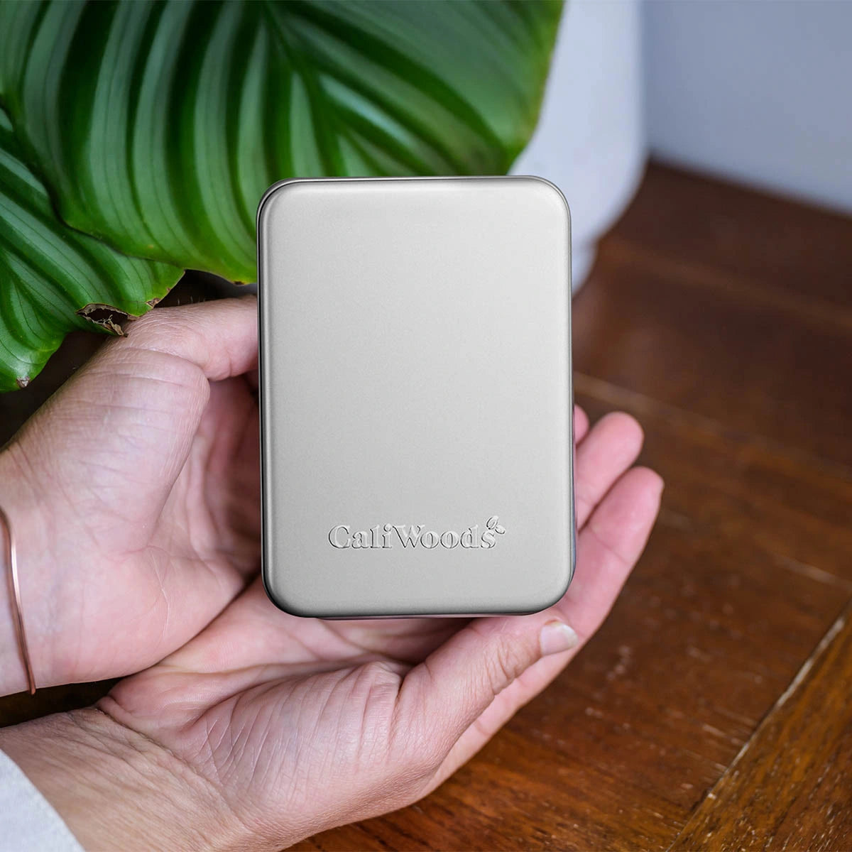 Hand holding a small metallic device with 'CaliWoods' branding, against a wooden surface and plant background.