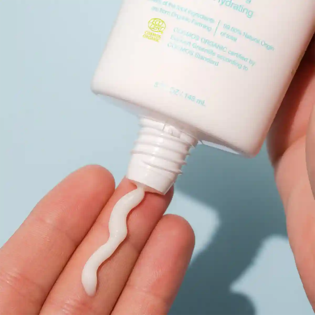 A close-up of a hand squeezing a white cream product tube onto another hand, showing a thick white cream being dispensed in a smooth smear against a soft light blue background.