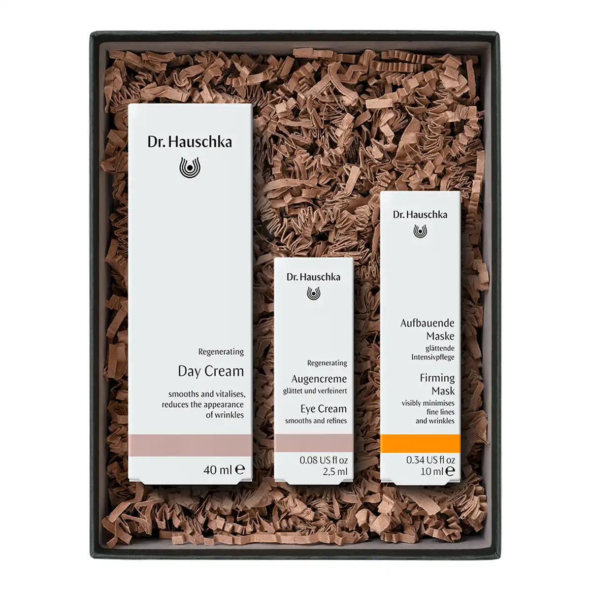 A Dr. Hauschka gift set containing three skincare products: Day Cream, Eye Cream, and Firming Mask, arranged in brown shredded paper inside a black box.