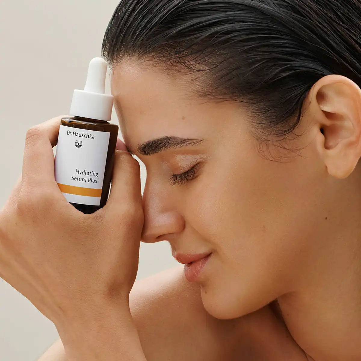 A close-up lifestyle shot of a woman holding a bottle of Dr. Hauschka Hydrating Serum Plus to her forehead, showing the product bottle against her skin.