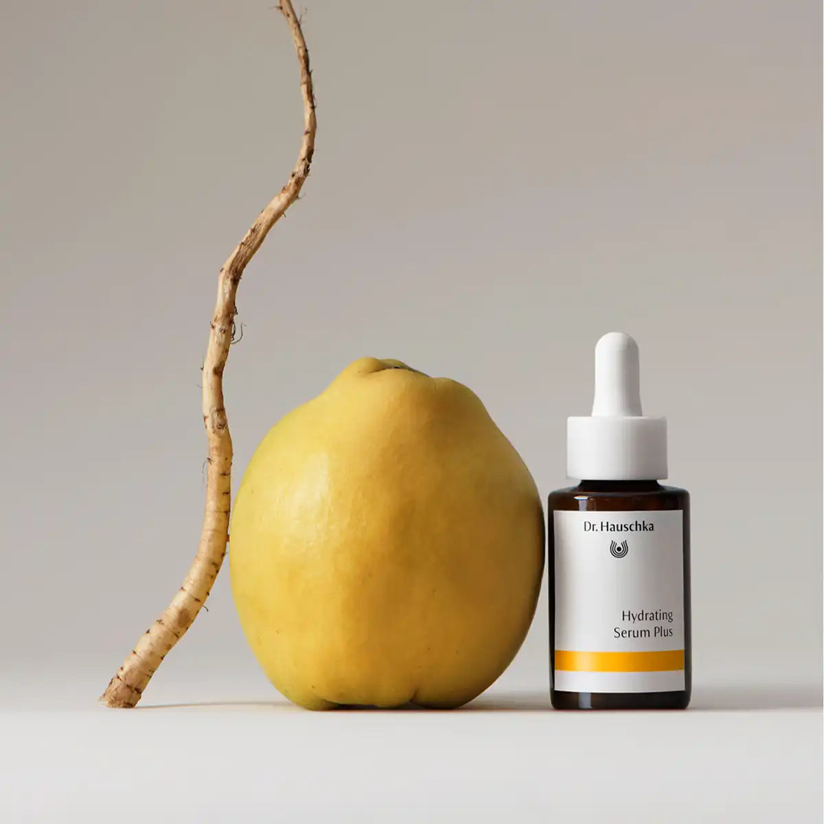 A small amber dropper bottle labeled Dr. Hauschka Hydrating Serum Plus stands beside a lemon and a long root on a clean light gray studio background.