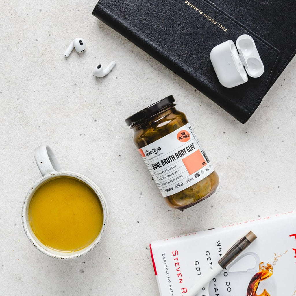 Jar of 'Body Glue' with a mug of tea, earbuds, and a book on a light surface.