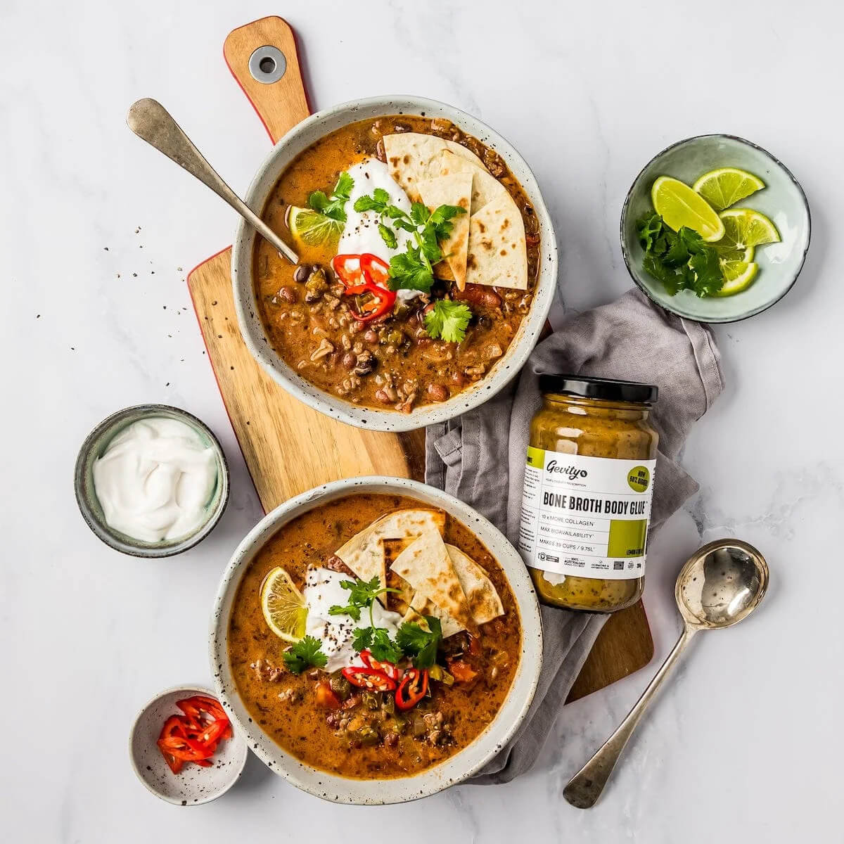 Two bowls of soup with tortilla chips, sour cream, and a jar of broth on a white surface.