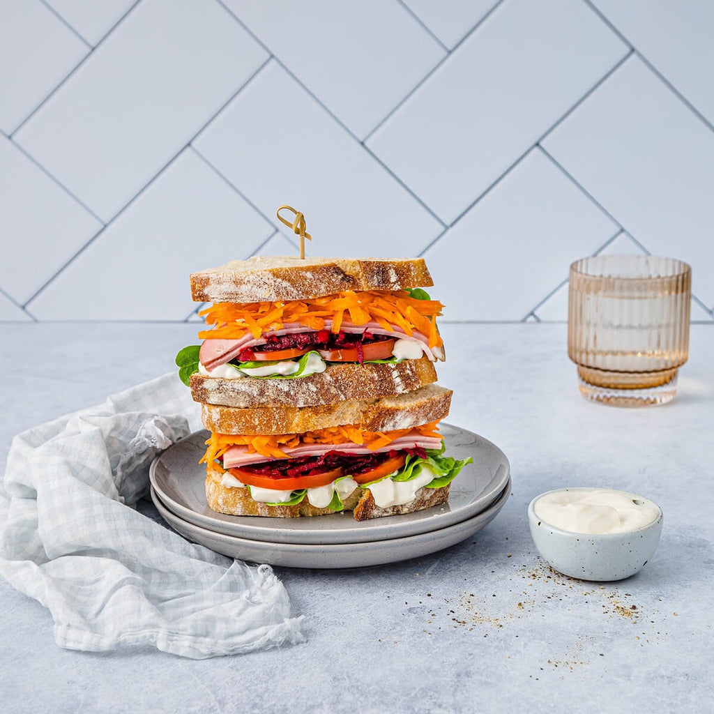 Stacked sandwich with various ingredients on a plate with a glass of water and small bowl.