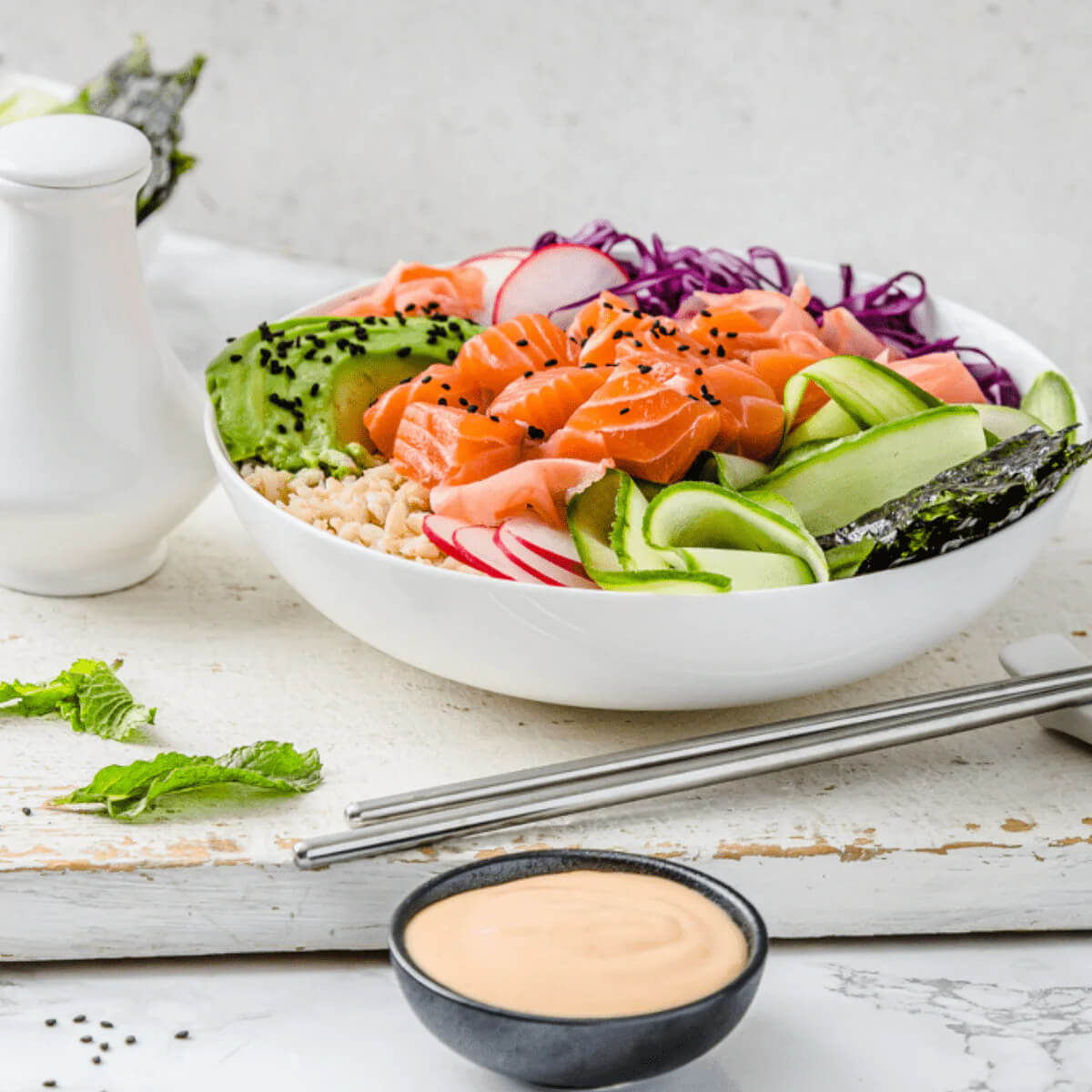 Salmon bowl with vegetables and a side of sauce on a wooden surface.