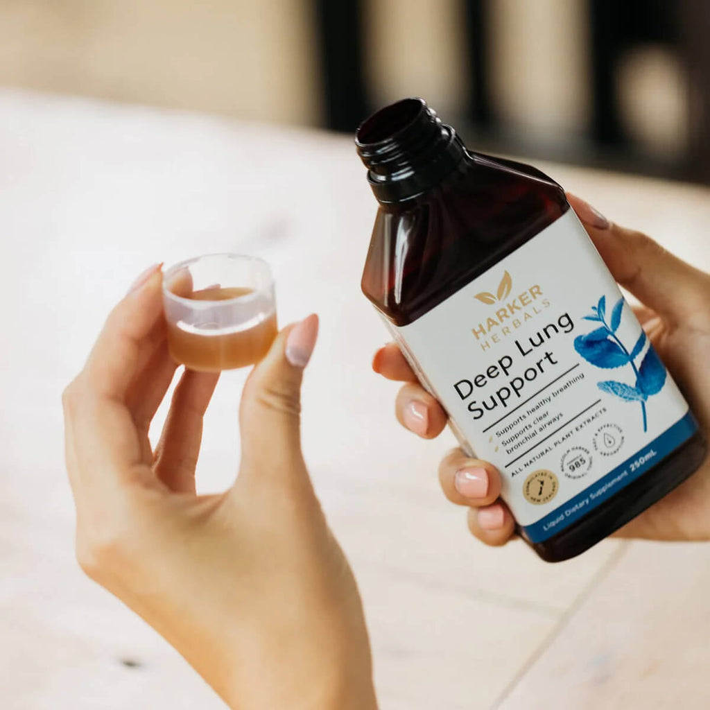 A person holds a dark amber bottle labelled 'Harker Herbs Deep Lung Support' beside a small, clear cup containing a light brown liquid, set against a softly blurred, light-coloured background.