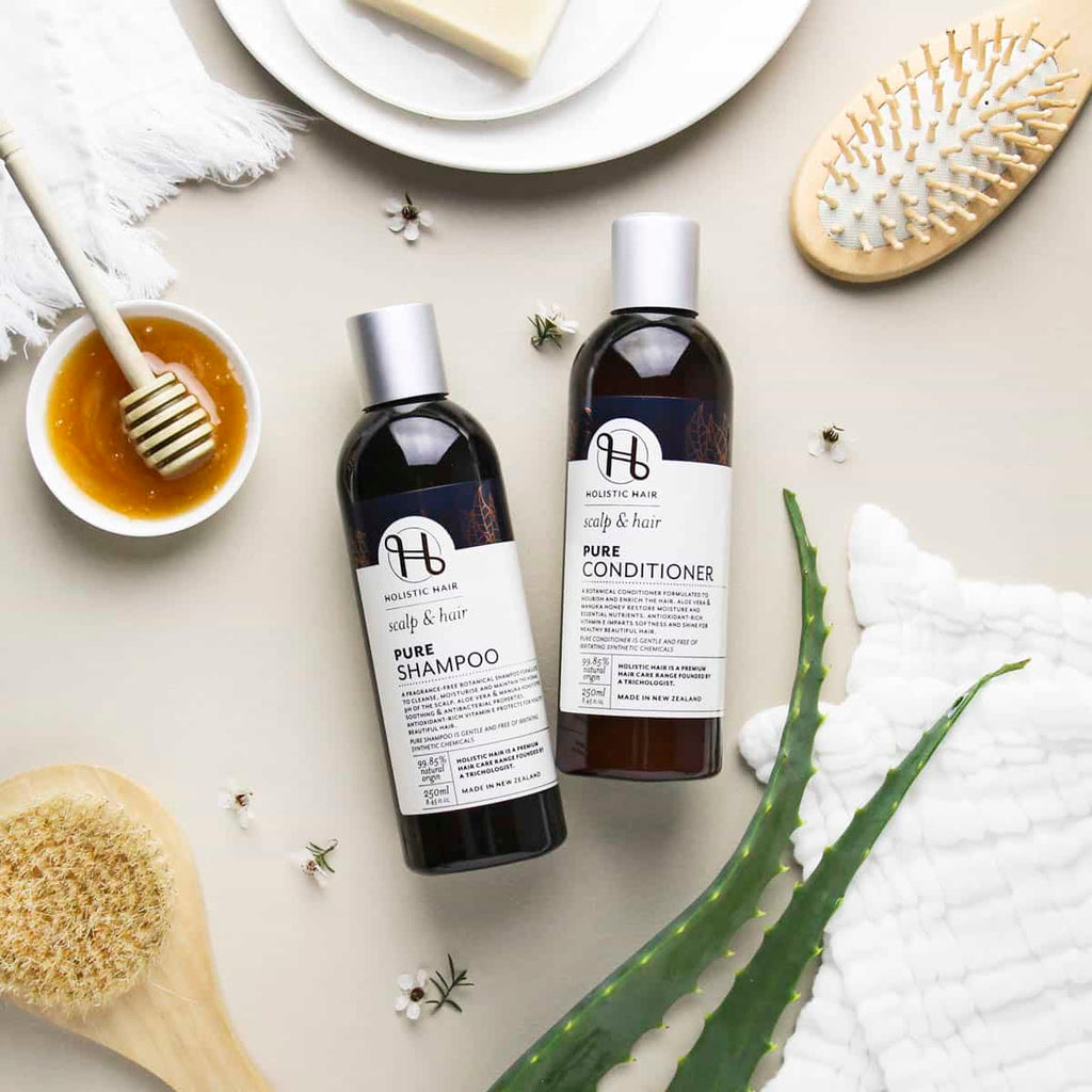 Two bottles of shampoo and conditioner with a bowl of honey, brush, and towel on a light surface.