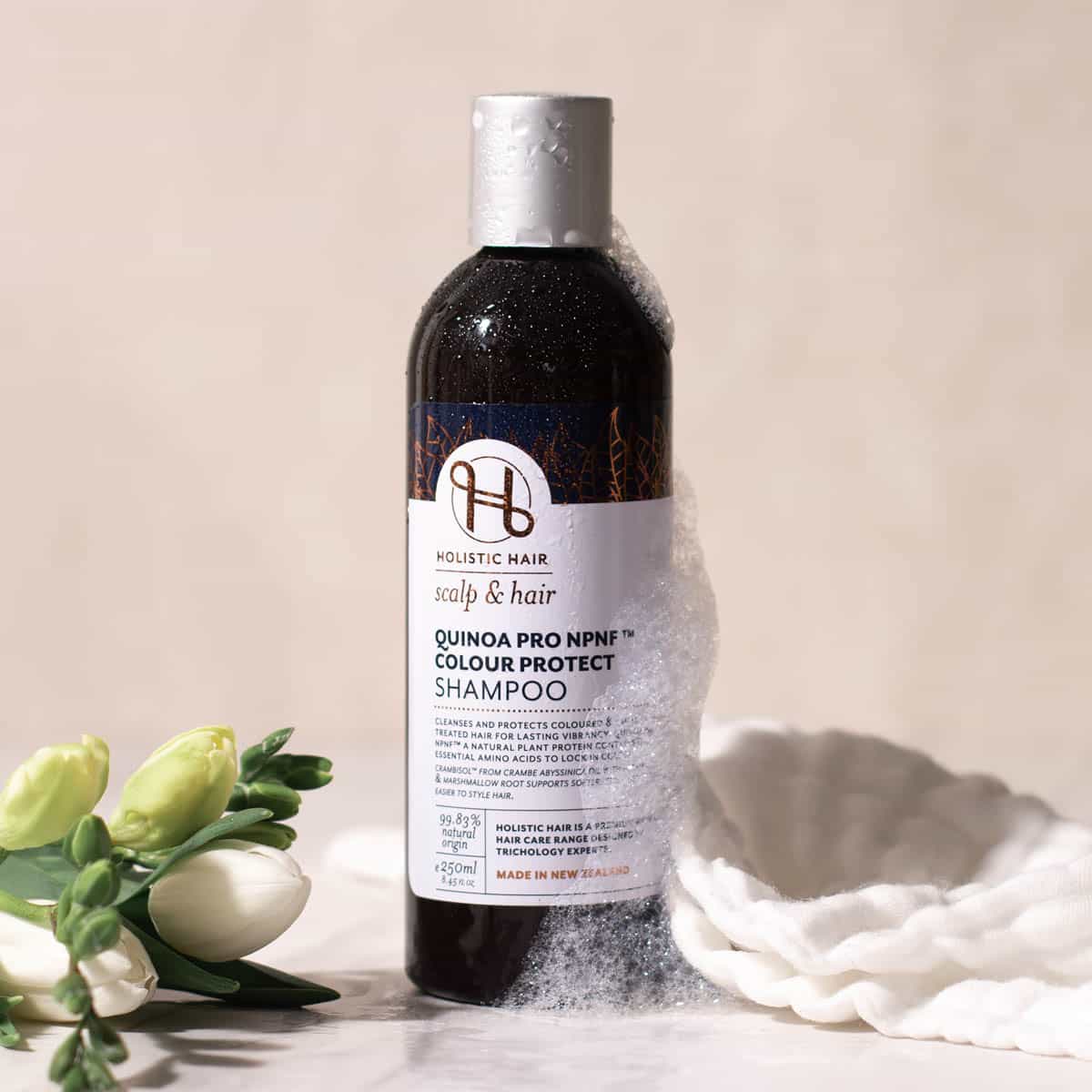 Bottle of Holistic Hair Colour Protect Shampoo with flowers and a white cloth on a beige background