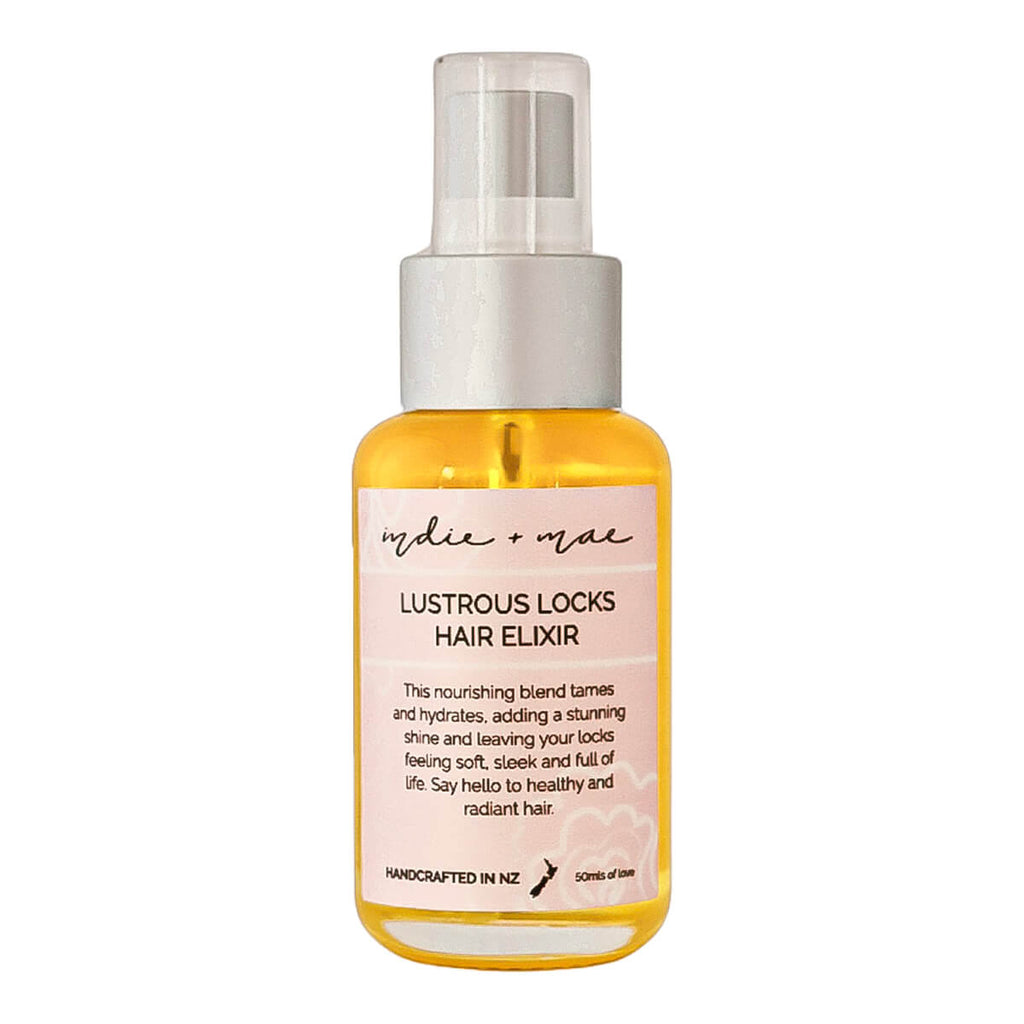 A bottle of Indie + Mae Lustrous Locks Hair Elixir, a golden yellow oil in a clear bottle with a white spray pump and a light pink label.