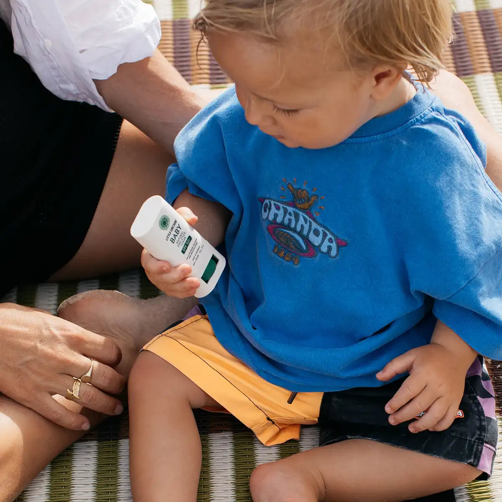 A toddler wearing a bright blue t-shirt and yellow shorts sits on a woven mat, holding a white tube of sunscreen.
