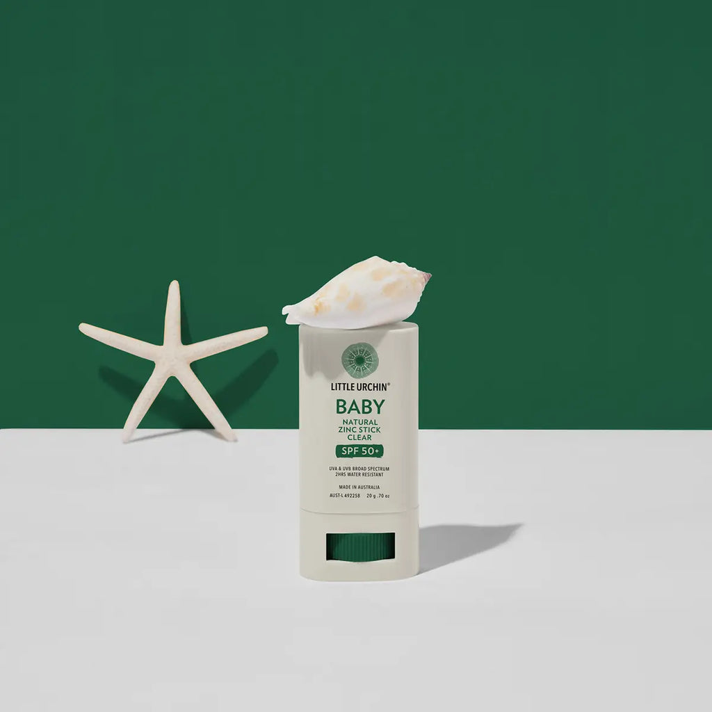 A baby sunscreen stick on a white surface against a dark green background, styled with a white seashell on top of the stick and a white starfish prop on the left.