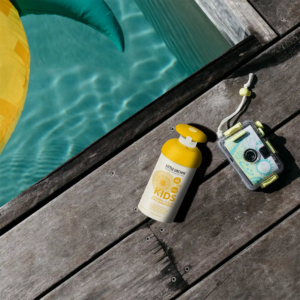 A bottle of sunscreen and a waterproof camera resting on a wooden deck next to a swimming pool.