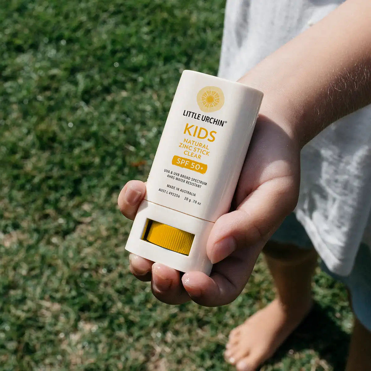 A hand holding a Little Urchin Kids Natural Zinc Stick SPF 50+ sunscreen stick outdoors on a grassy lawn.