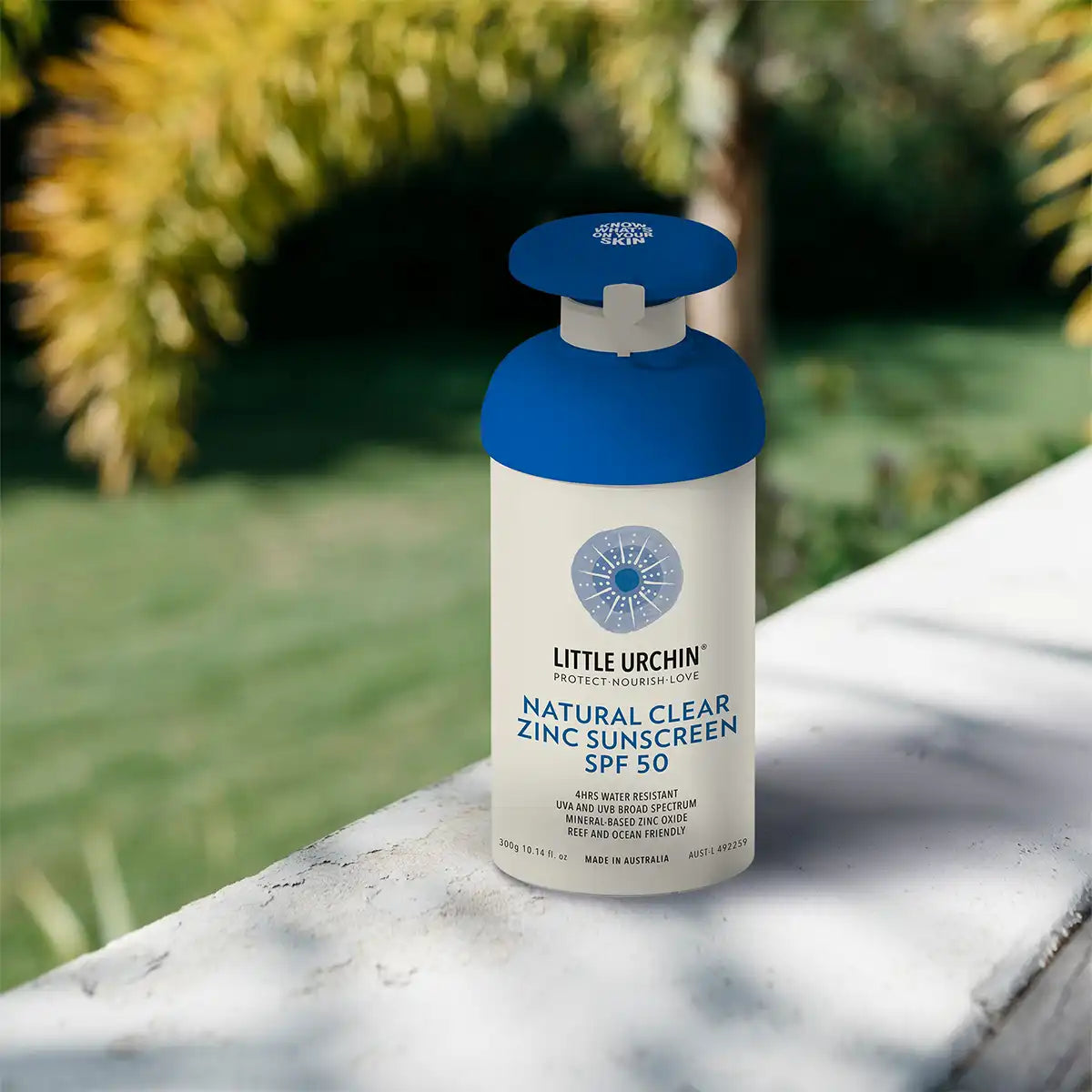 Little Urchin Natural Clear Zinc Sunscreen SPF 50 pump bottle on a white stone ledge with palm leaves behind it in warm sunlight
