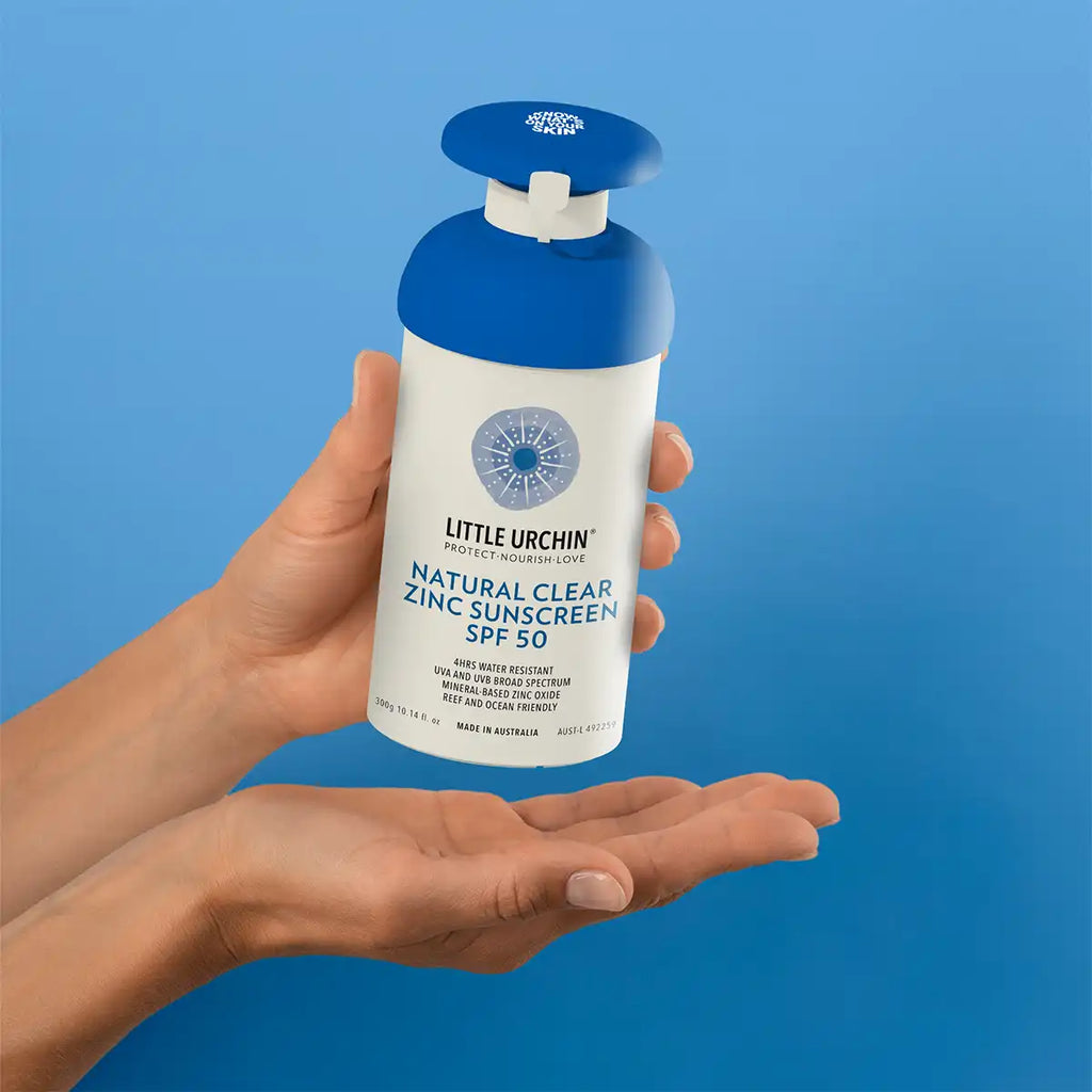 Two hands hold a Little Urchin Natural Clear Zinc Sunscreen SPF 50 pump bottle with a blue pump head against a bright blue background