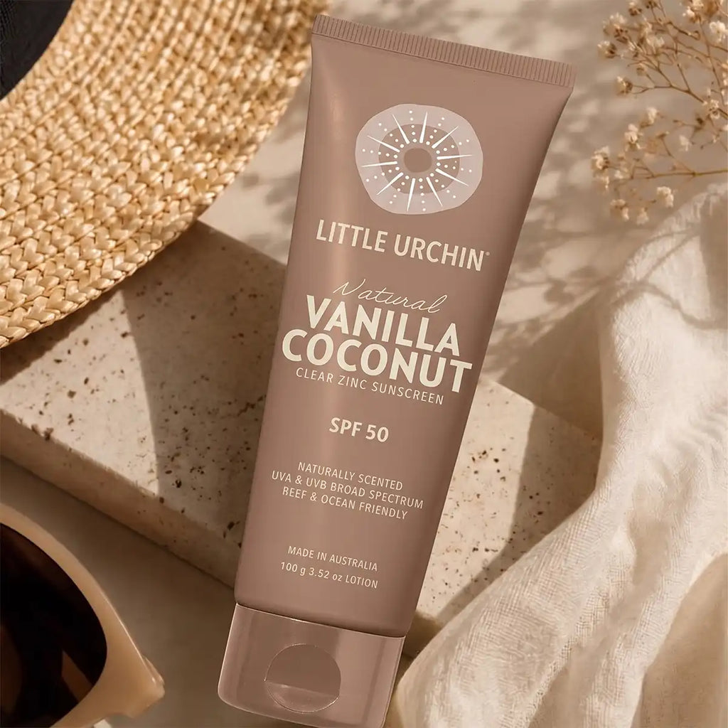 A tube of Little Urchin Natural Vanilla Coconut Clear Zinc SPF 50 sunscreen, styled in a flat-lay arrangement with a woven straw hat, a speckled stone slab, a white fabric cloth, and dried flowers.