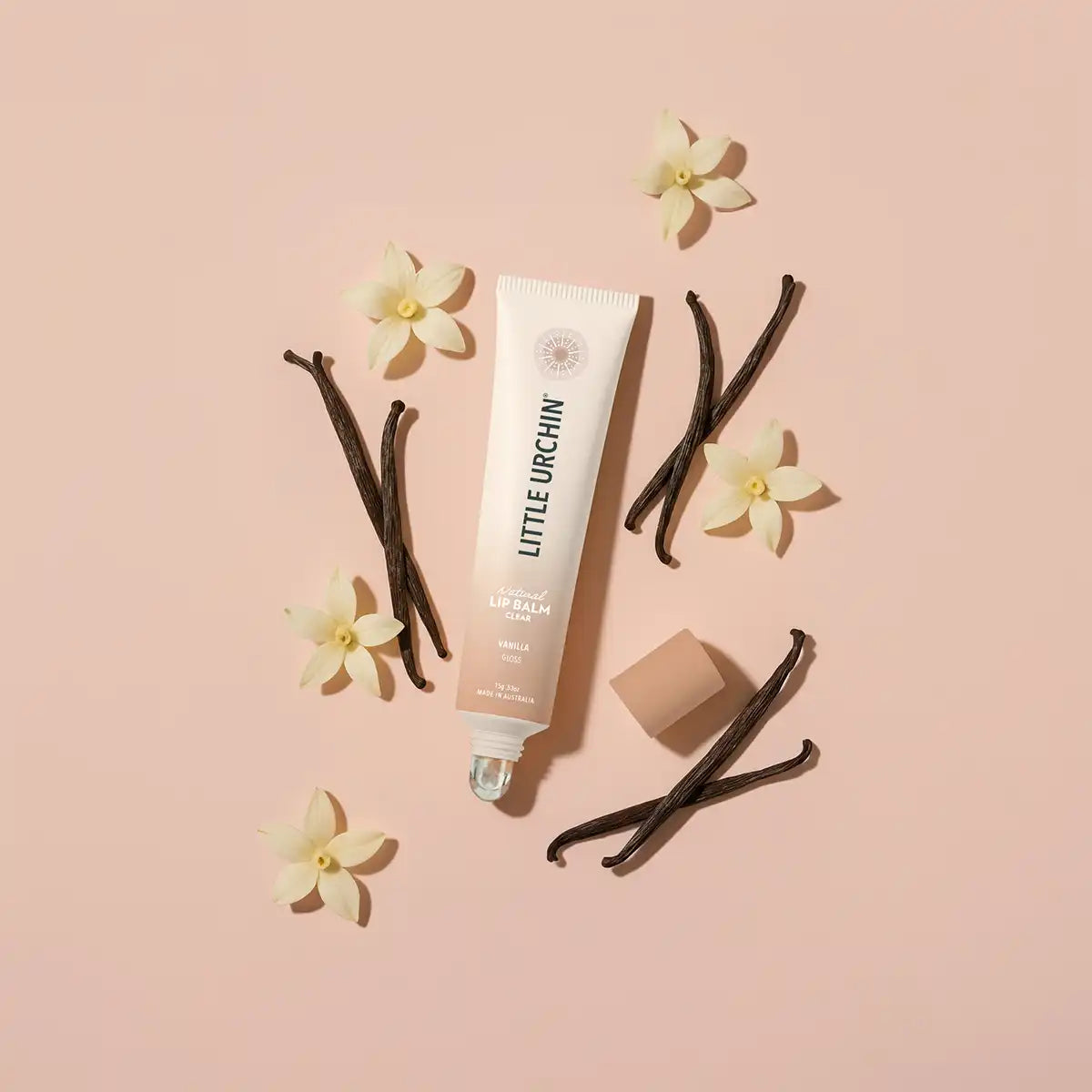 A tube of Little Urchin Vanilla lip balm is arranged on a soft pink background with vanilla beans, small white flowers, and the cap placed beside it.