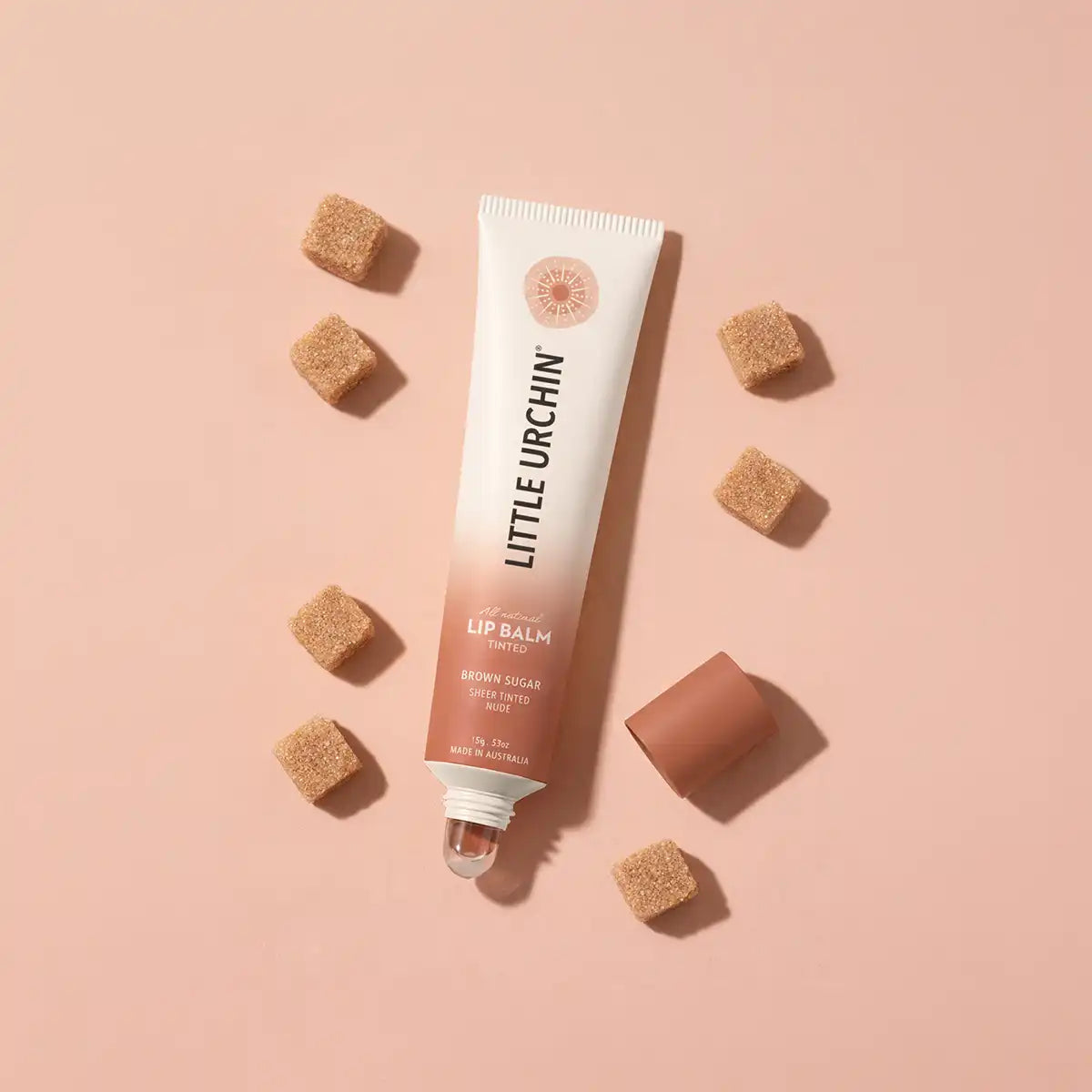 A tube of Little Urchin Brown Sugar Lip Balm is displayed on a dusty pink background, surrounded by scattered brown sugar cubes.