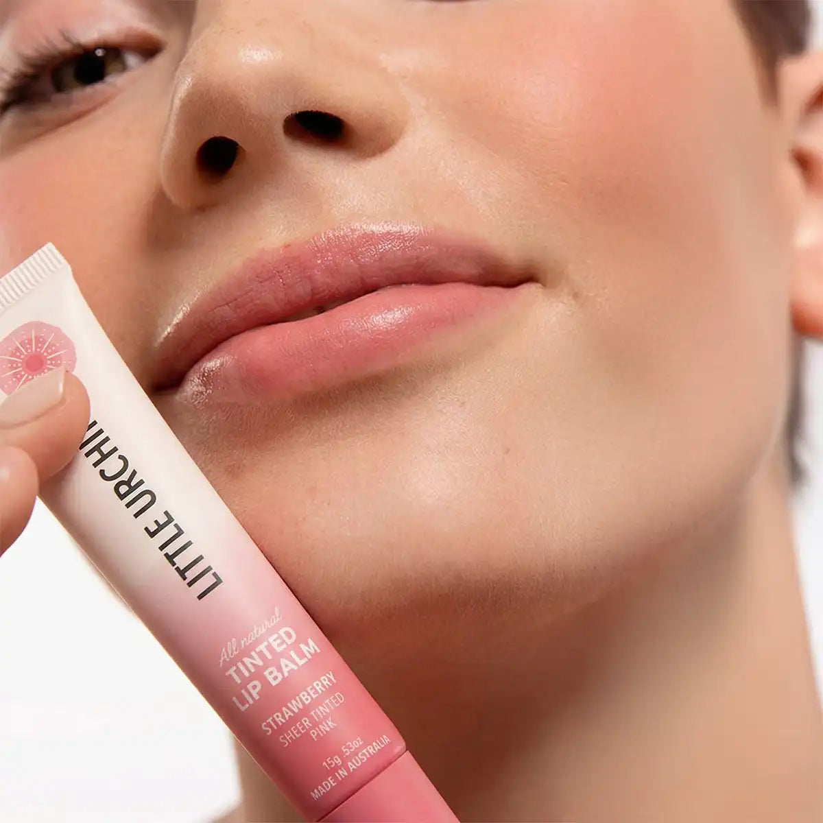 Close-up of a person holding a tube of Little Urchin Tinted Lip Balm in Strawberry, with the product tube shown against the model's lips and cheek.