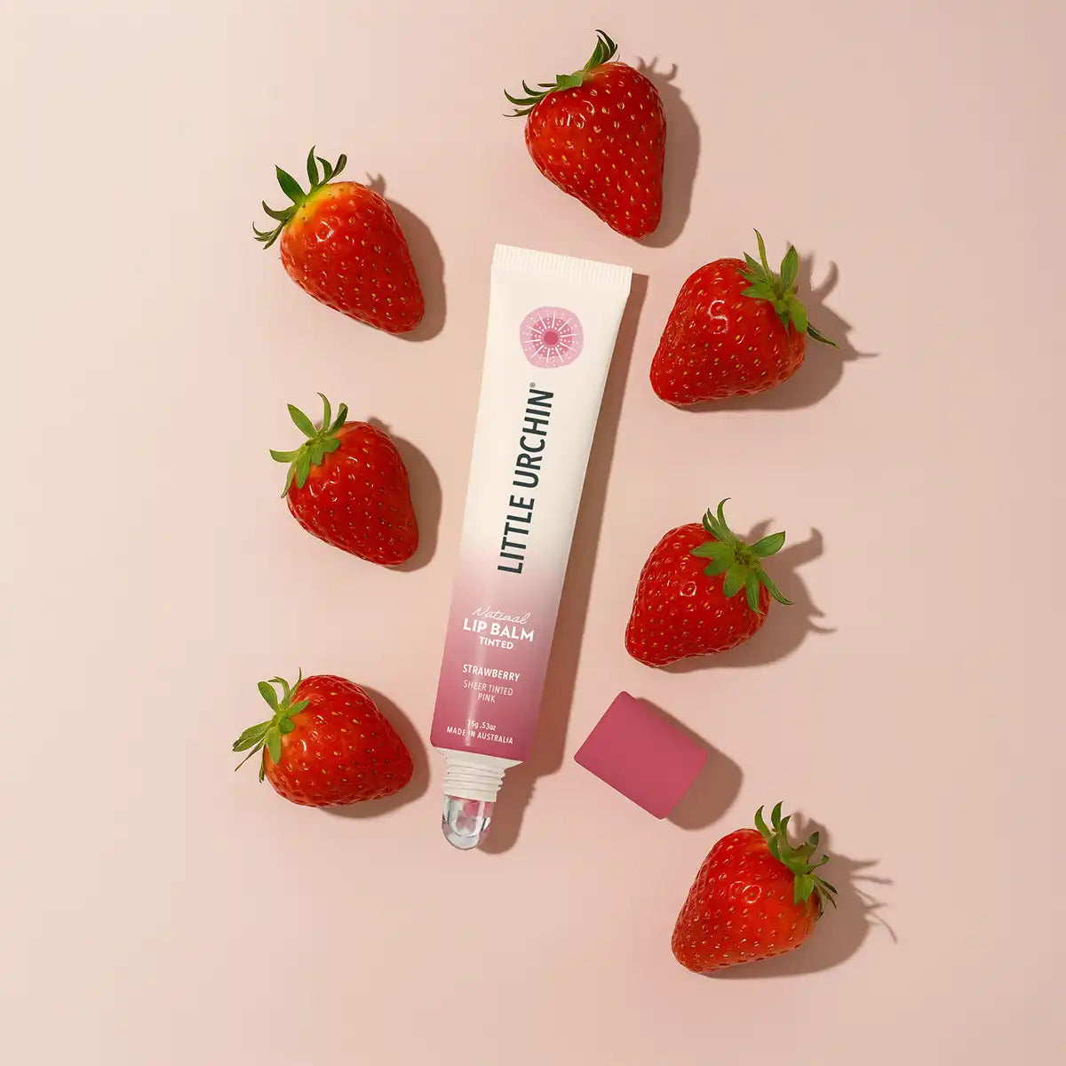 A tube of Little Urchin Strawberry Lip Balm is arranged on a light pink background, surrounded by fresh strawberries.