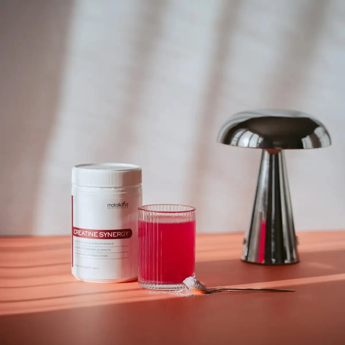 A white tub of Creatine Synergy powder stands next to a glass of bright pinkish-red liquid and a small spoonful of powder on a terracotta surface, with a metallic mushroom-shaped lamp in the background.