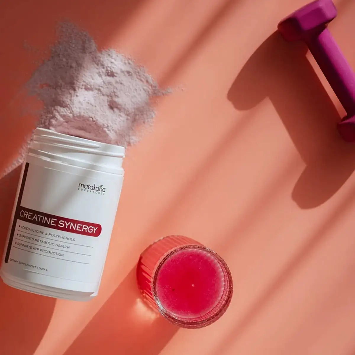 A white supplement jar labeled Creatine Synergy with pink powder spilling out, styled on a peach background with a pink dumbbell and a pink drink in the foreground.