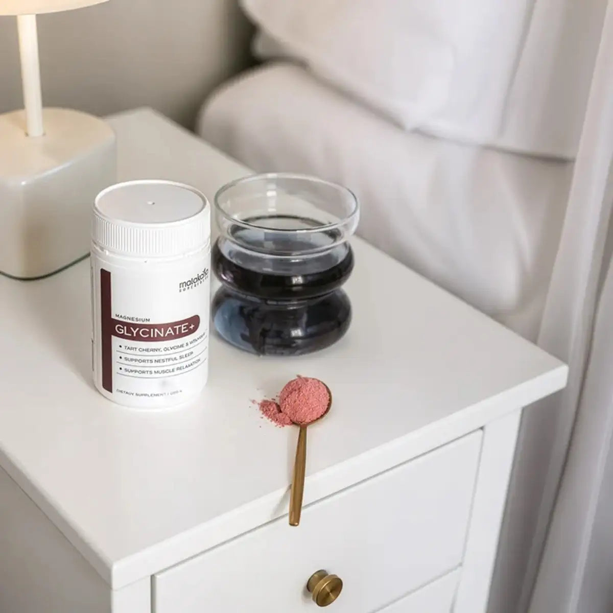 A white supplement bottle and a glass of dark powder sit on a white bedside table, with a small spoonful of pink powder nearby in a softly lit bedroom setting.