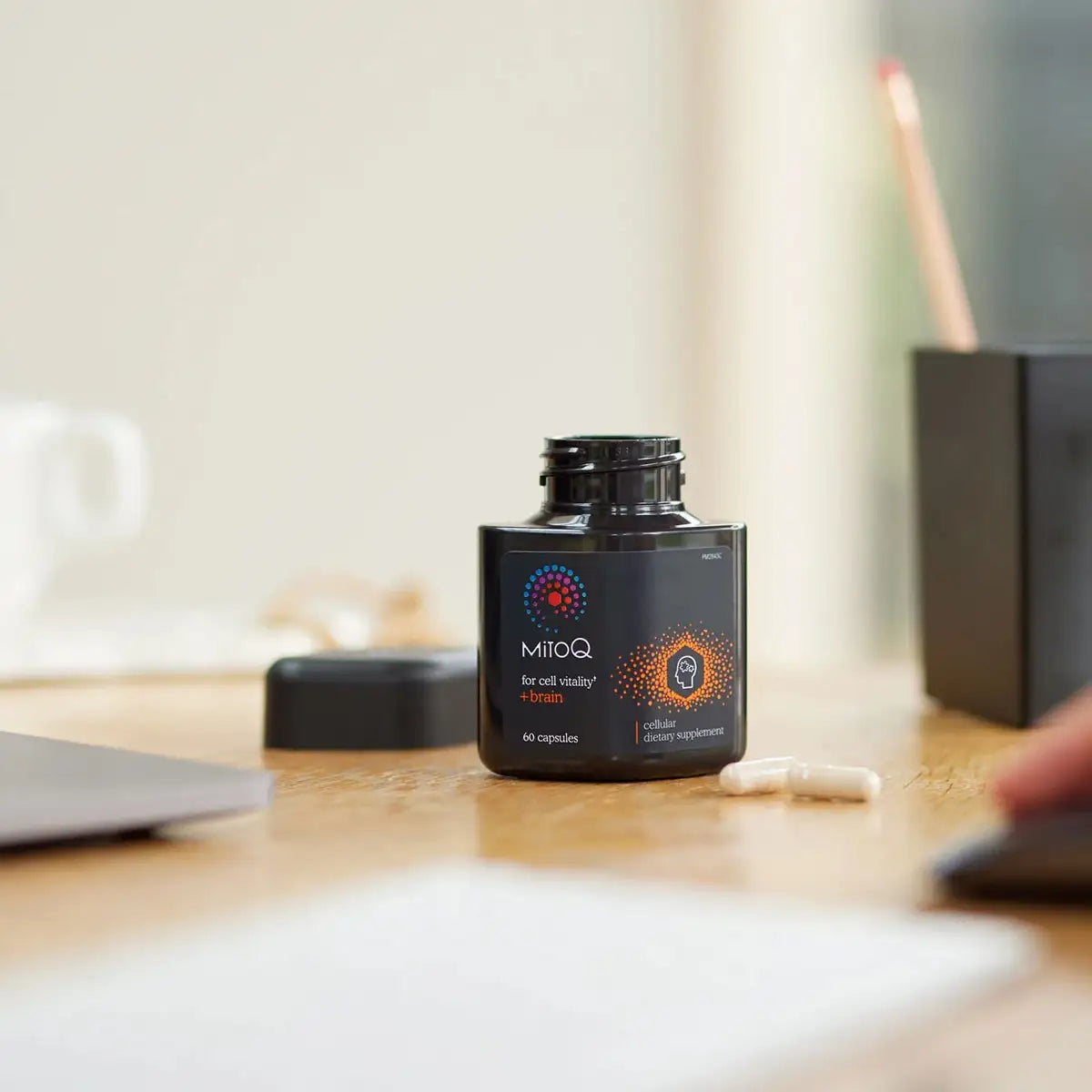 A black bottle of MitoQ capsules sits on a wooden desk with its cap off, alongside a few white capsules.
