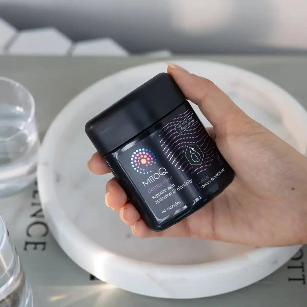 A hand holding a black supplement jar labeled MitoQ Derma Elite against a marble tray and blurred background.