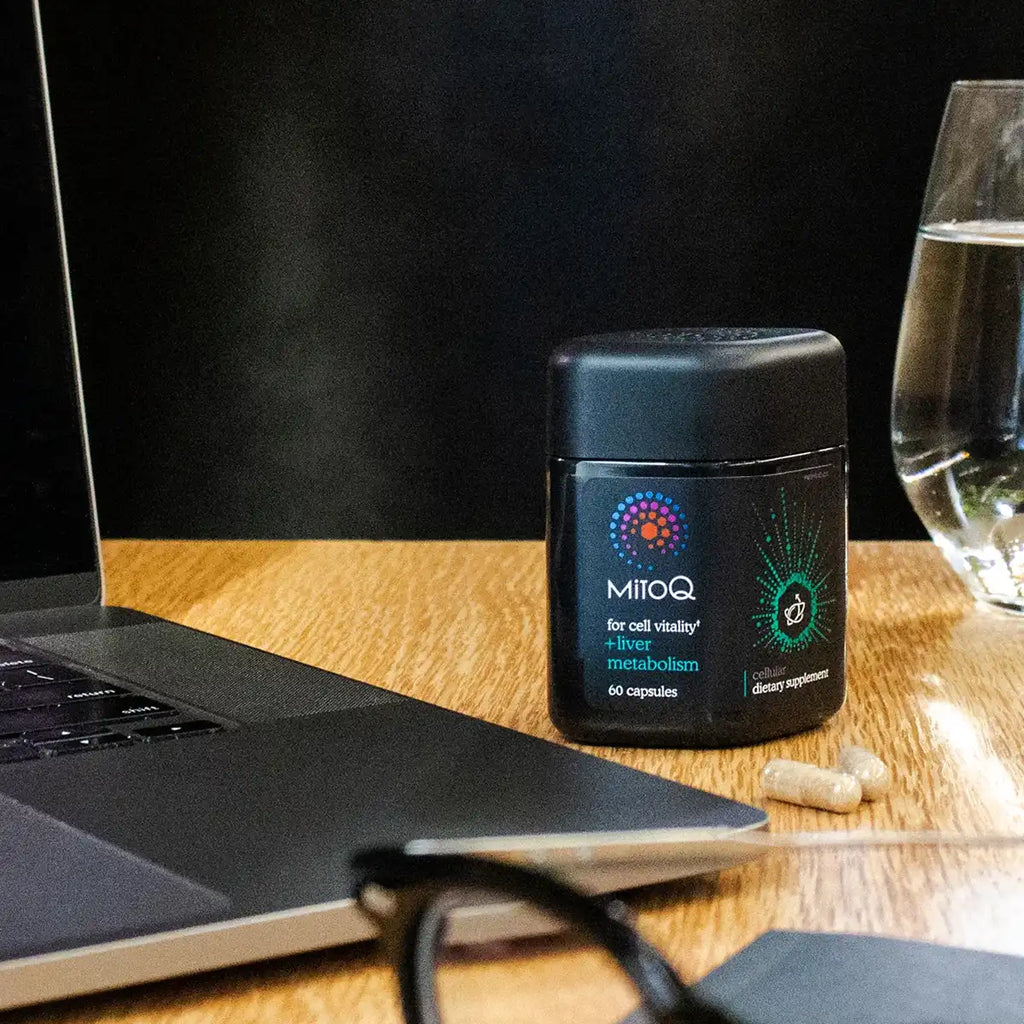 A black bottle of MitoQ dietary supplement capsules sits on a wooden desk next to a laptop, a glass of water, and eyeglasses.