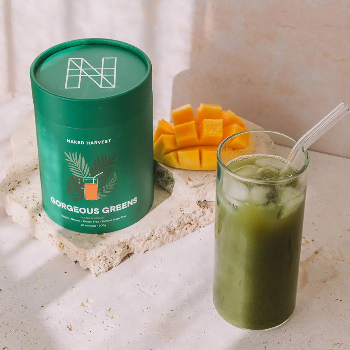 Green smoothie in a glass with a straw, next to a container labeled 'Naked Harvest' and sliced mangoes on a light surface.