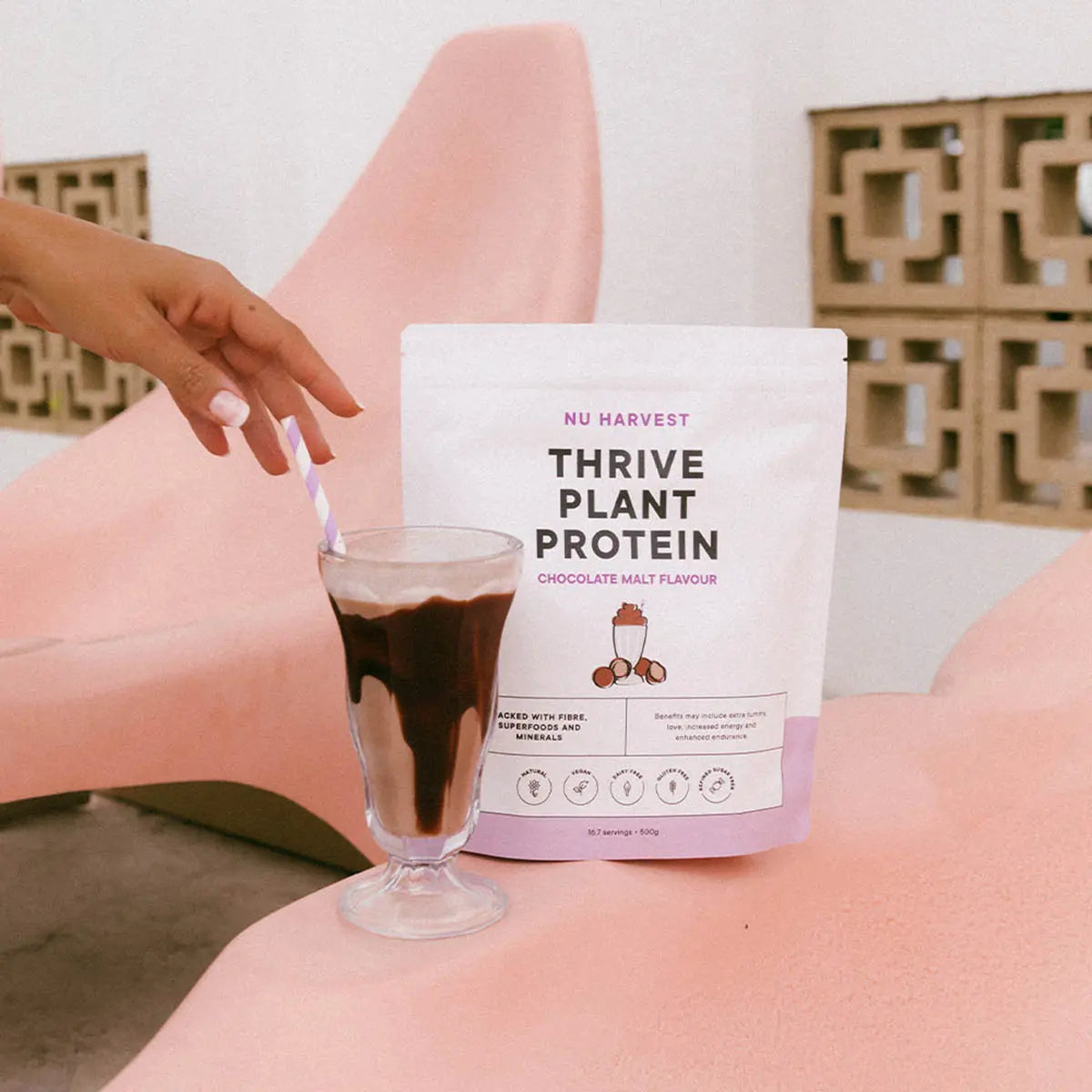 Person holding a glass of chocolate milk next to a package of Nu Harvest Thrive Plant Protein.