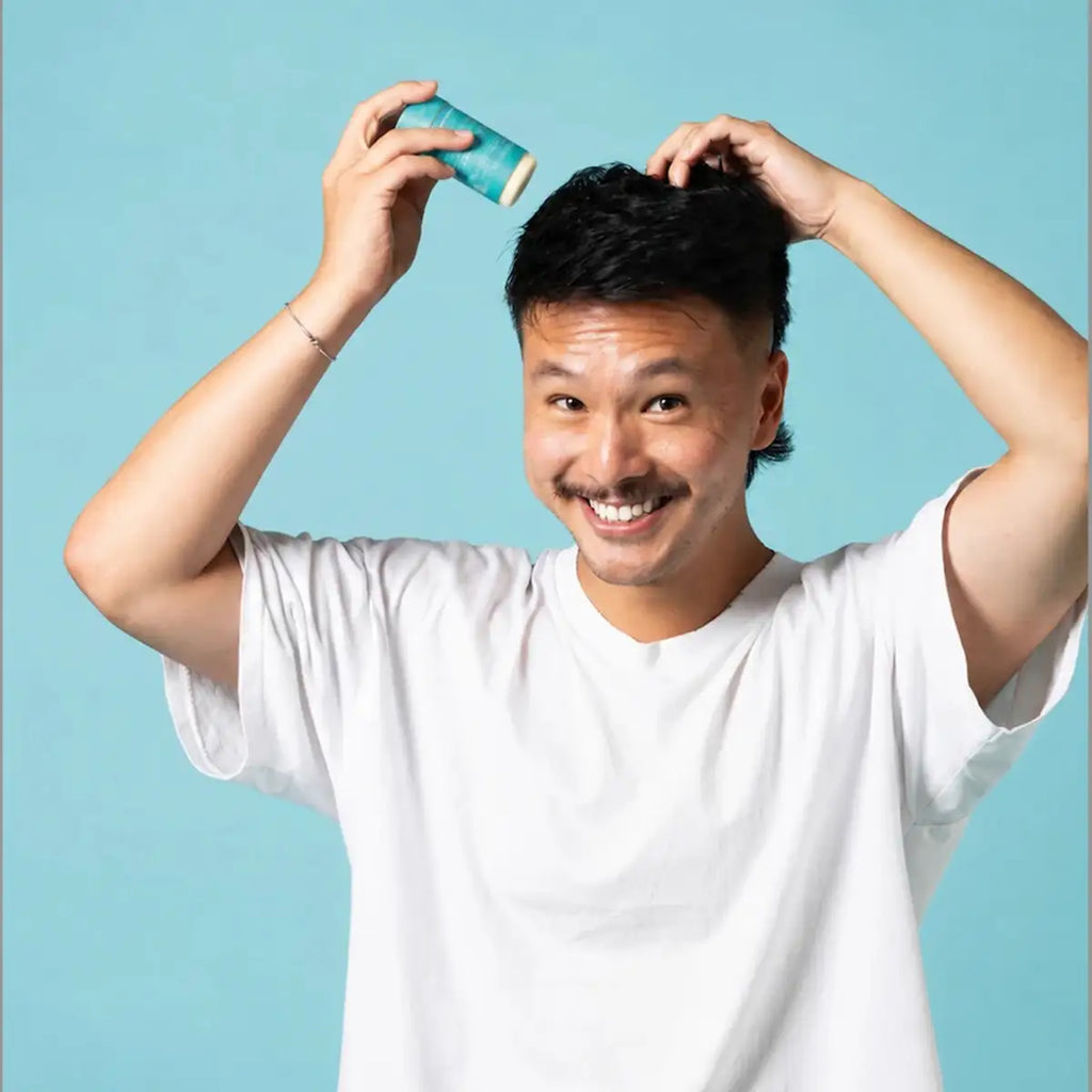 A smiling man in a white t-shirt holds a slo hair styling wax stick to his hair and looks at the camera against a bright teal background