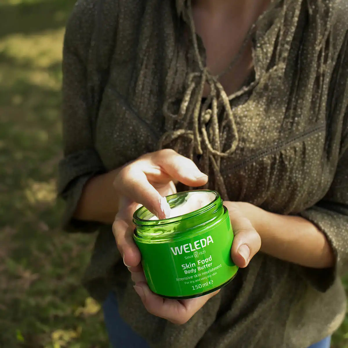 Person holding jar of Weleda Skin Food Body Butter and scooping out some onto their finger. In an outdoor setting with dappled light.