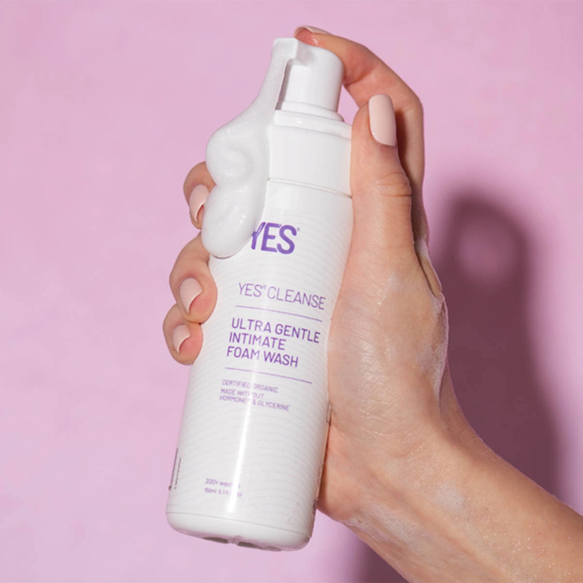 A hand holding a white bottle of YES Cleanse Ultra Gentle Intimate Foam Wash, with white foam visible on the bottle and dripping from the dispenser, against a solid pink background.